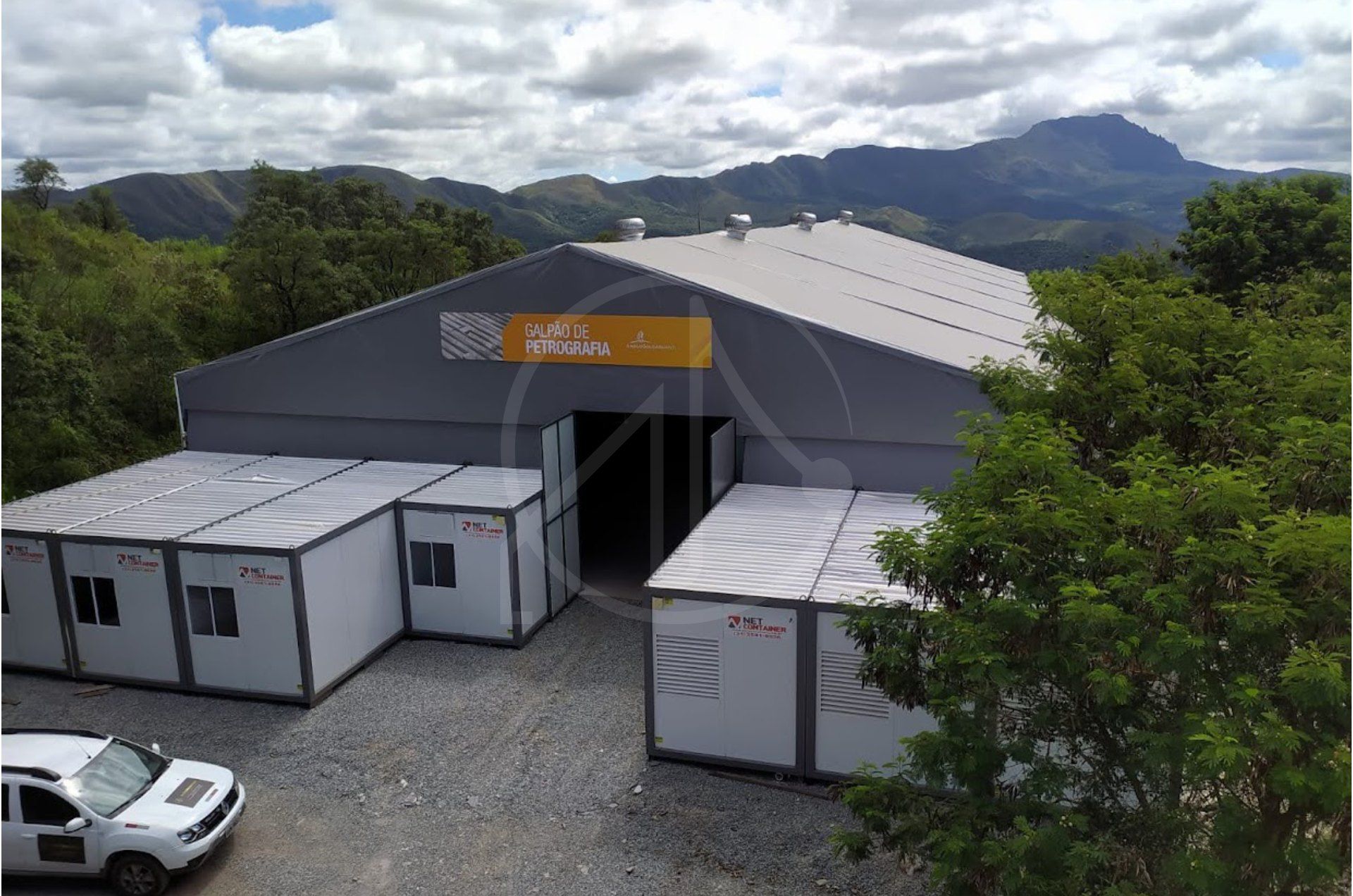 Galpão temporário cinza com contêineres de escritório ao lado, cercado por montanhas